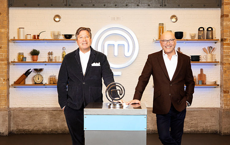 John Torode and Gregg Wallace stand either side of the Celebrity Masterchef 2022 trophy on a countertop both wearing white shirts and black jackets and trousers smiling at the camera, the Masterchef logo in the background