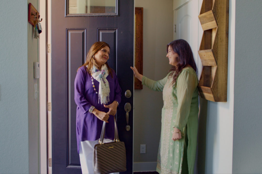 Sima Taparia is welcomed into Rupam Kaur's home. Sima wears a purple top and holds a brown handbag while Rupam wears a light green dress with her brunette hair worn down