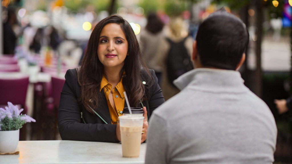 Aparna from Indian Matchmaking sits opposite her date at a table wearing a mustard shirt and black leather jacket