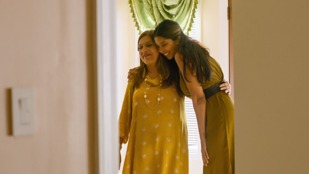 Nadia, wearing an olive green dress leans over and hugs Sima from the side who wears a yellow dress and a gold necklace, both have their eyes closed