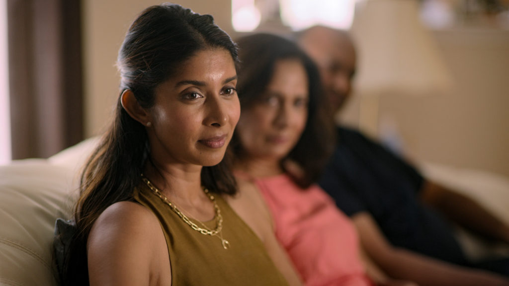 Indian Matchmaking's Nadia sits on a sofa with her parents in the background in soft focus, she wears a brown top with two gold chains and gazes across to the right of the camera