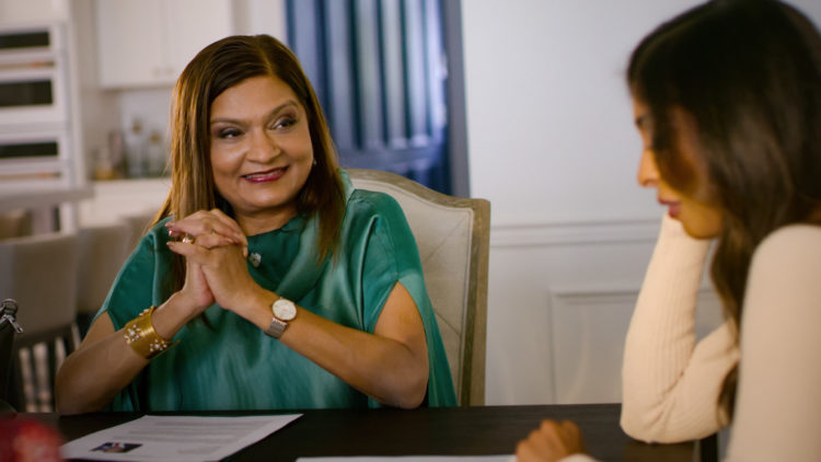 Indian Matchmaking's Sima sits adjacent to a client at a table with her elbows on the table and her hands clasped together, she's smiling wearing a silk turquoise top