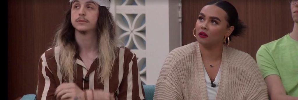 Turner and Jasmine on BB24 sit and wait to hear who is being evicted. Turner wears a brown and cream vertically striped shirt, Jasmine wears red lipstick and a cream knitted cardigan, a white t shirt and gold hoop earrings.