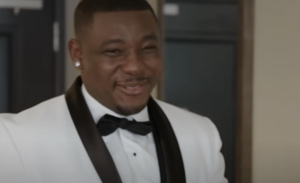90 Day Fiancé's Kobe smiles wearing a white suit and a black tow tie with a diamond earring