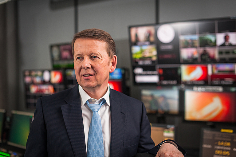 Bill Turnbull wears blue tie and black blazer with white shirt in front of TV screens.