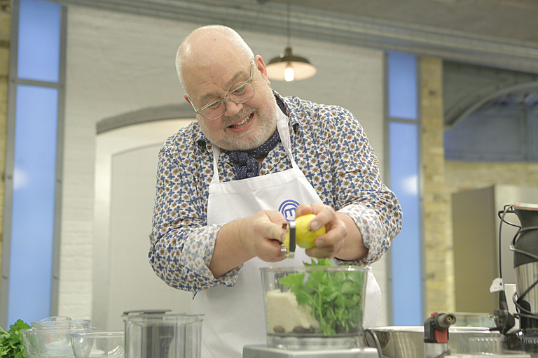 Cliff Parisi grates lemon peel into a food processor on Celebrity Masterchef wearing a patterned shirt, apron and glasses