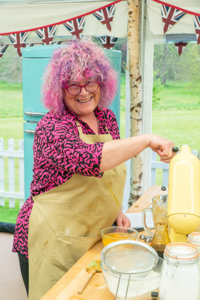 Carole baking in the tent with pink hair, pink shirt and beige apron