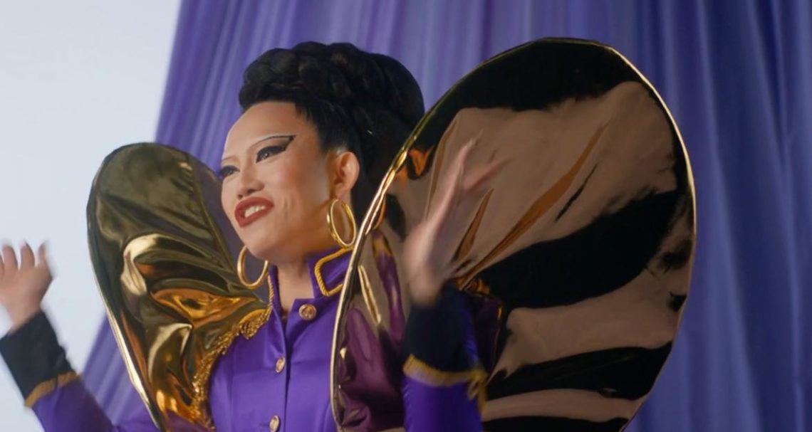 Le Fil wears purple wing outfit on RuPaul's Drag Race UK and holds hands out.