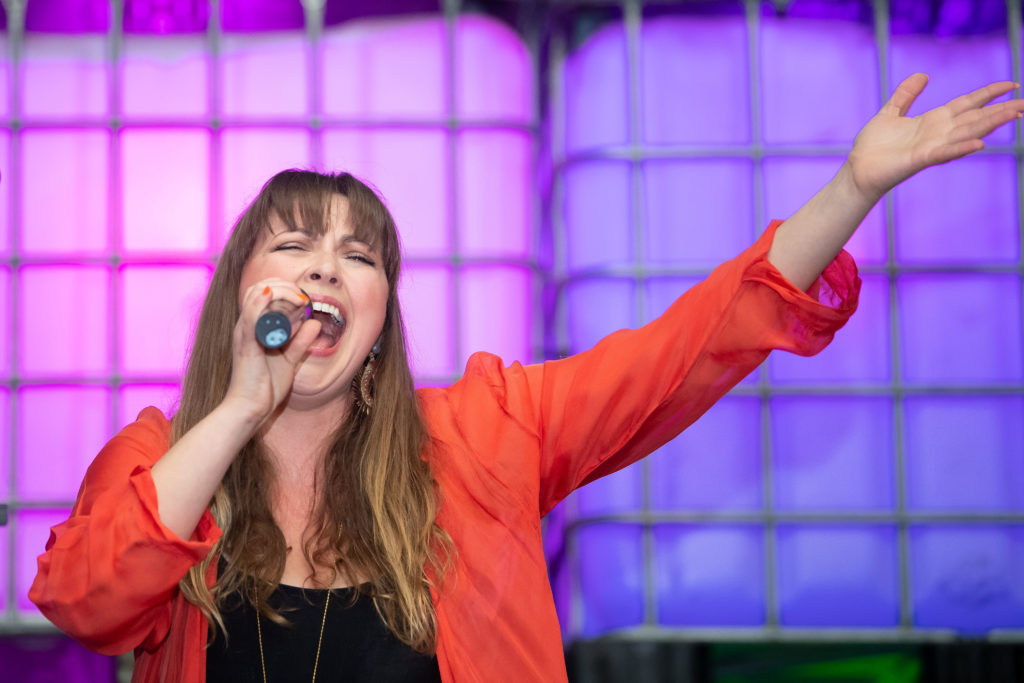 Charlotte Church sings into microphone wearing red blazer and black top holding left arm in the air