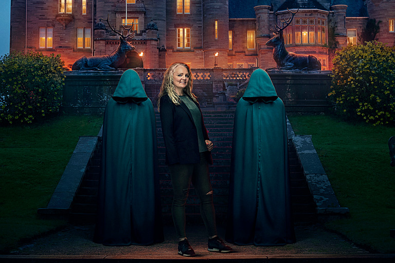 Nicky shows right side of full body next to two green cloaks with castle in background.