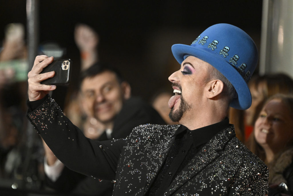 Boy George sticks tongue out and holds phone to his face for selfie wearing glitter black suit jacket and black top and a blue hat