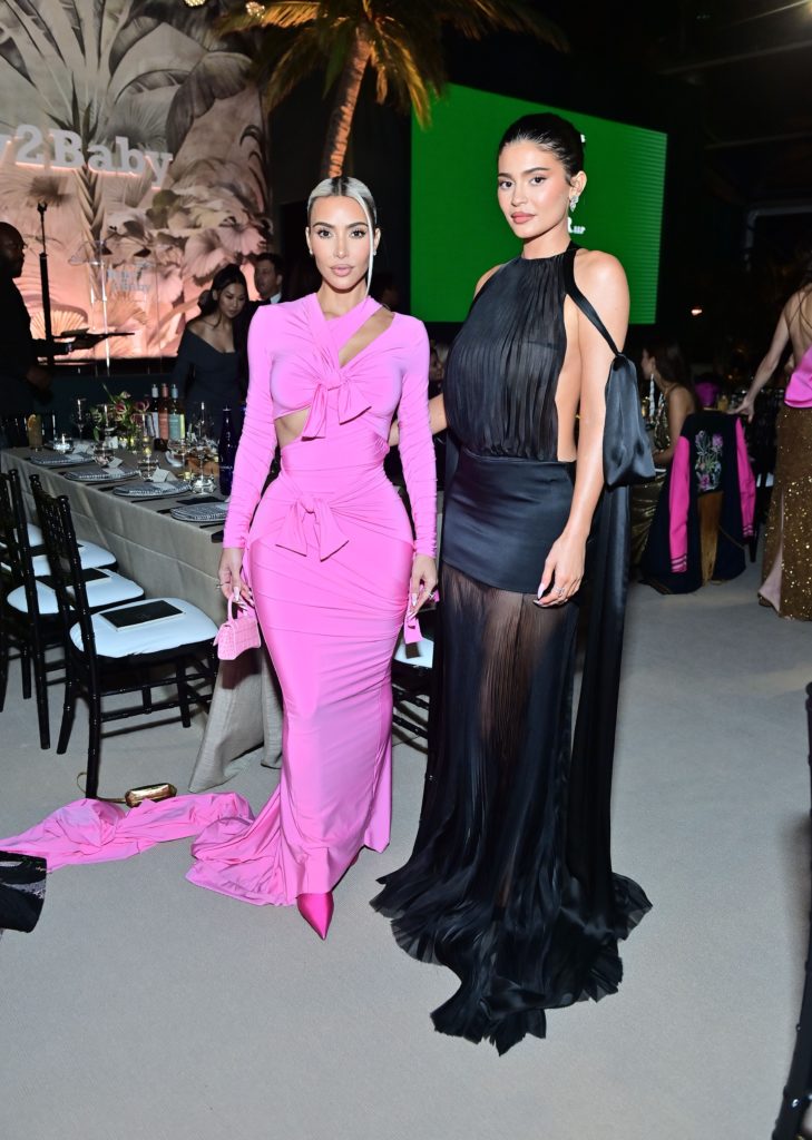 Kim Kardashian poses in a pink dress with a long train and sleeves standing next to Kylie Jenner who wears a long black sleeveless dress