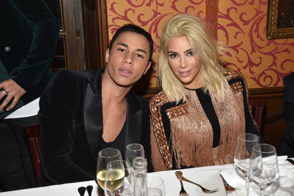Olivier Rousteing and Kim Kardashian sit together at a table, with orange wallpaper behind them, Kim has blonde hair and a copper coloured jacket and Olivier wears a black low cut top and blazer