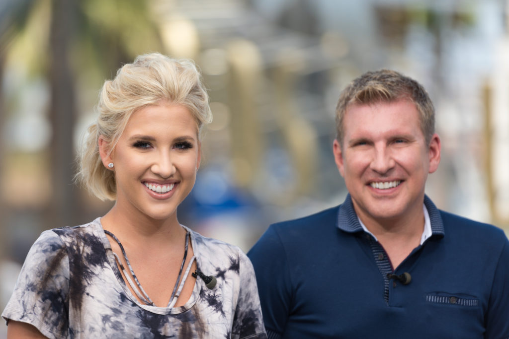 Todd And Savannah Chrisley stand next to one another smiling at the camera, Savannah wears a tie dye top and Todd wears a navy polo neck top