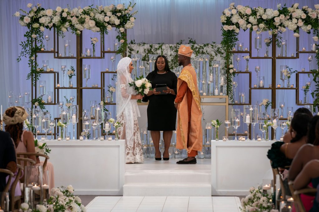 SK Alagbada wears orange headpiece and robe while Raven Ross wears white veil and wedding dress while holding white roses at wedding altar. White roses surround them. Priest stands inbetween them while family members sit in chairs watching.