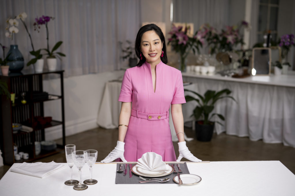 Sara Jane Ho wears pink dress and white gloves with hands on dinner table with plates and glasses. Plants in background.