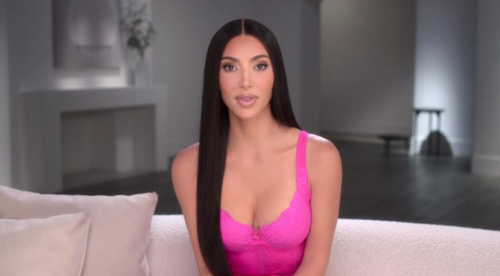 Kim Kardashian wears a pink lace top as she sits on a white sofa and stares into the camera