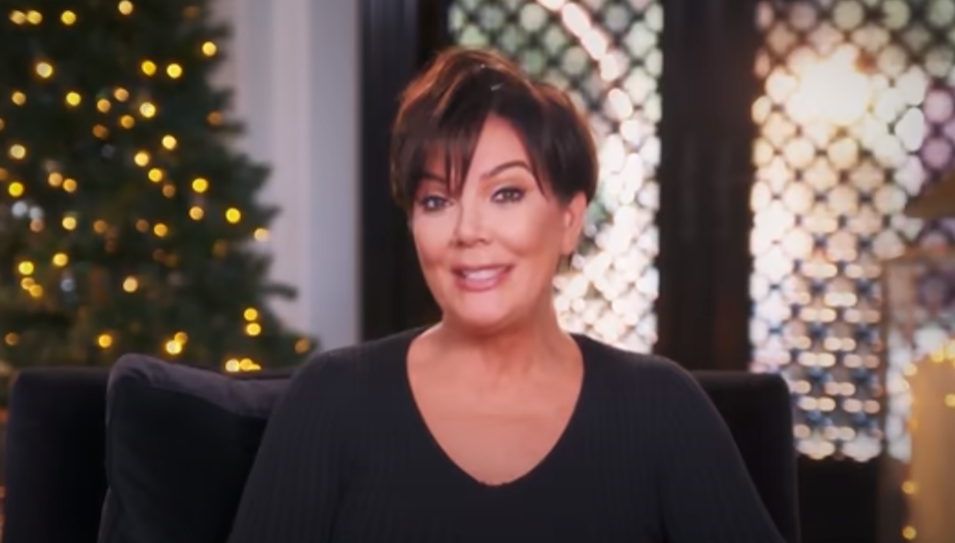 Kris Jenner wears a black top and sits in front of a green Christmas tree with lights