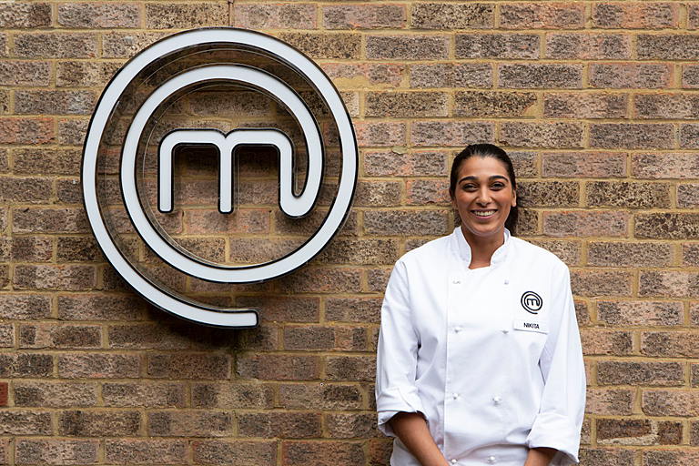 Nikita stands in chef whites next to MasterChef sign