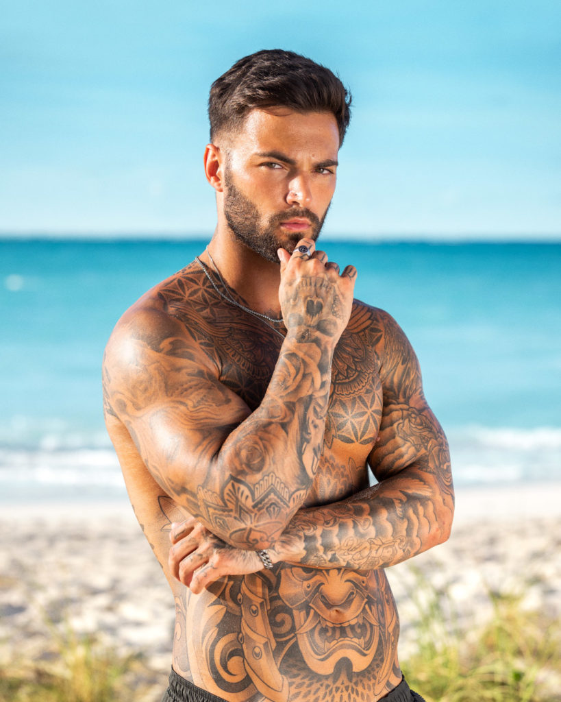 Ethan Smith holds right hand to chin in front of beach background. Covered in tattoos.