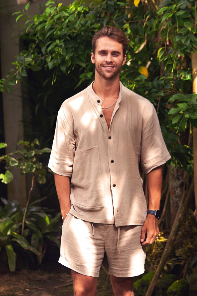 Julius Cowdrey smiles in Bali wearing beige co-ord shirt and short with greenery in background