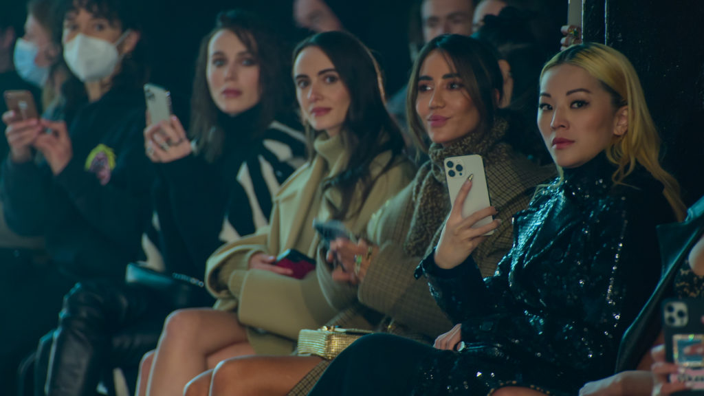Tina Leung attends fashion show and sits next to other women holding her phone up next to the catwalk