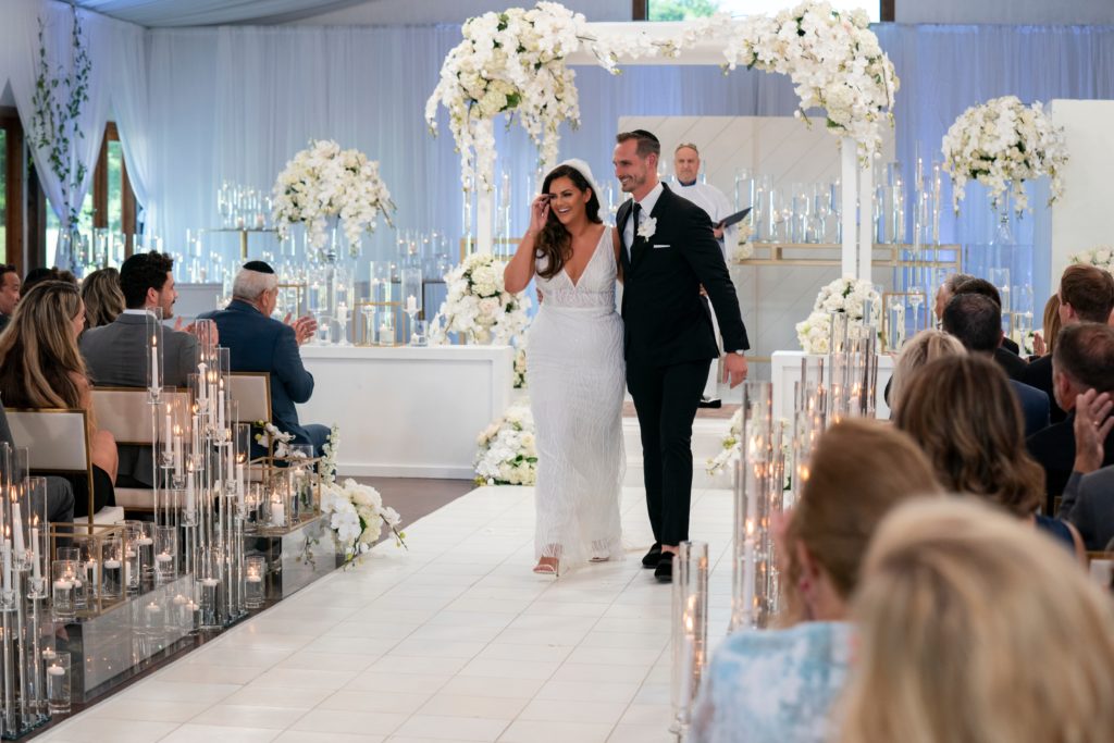 Alexa Lemieux in white long dress and Brennon Lemieux in black suit walk on white tiles with floral arch behind them, people clapping nearby.