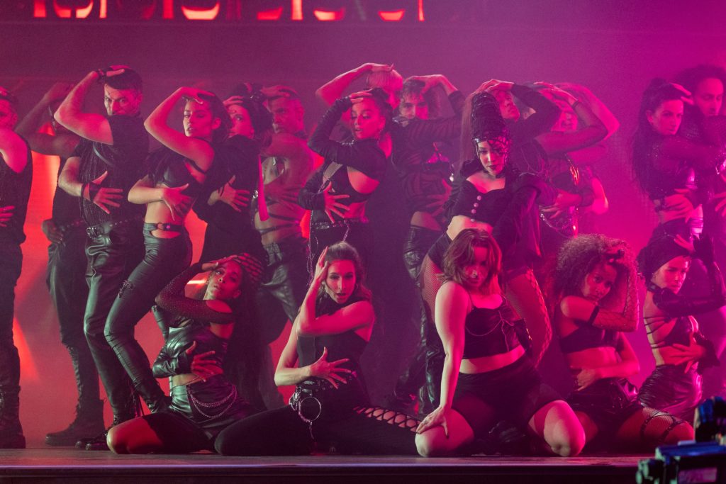 Brandi Chun and a group of dancers dressed in black perform on the Dance 100 stage which has been lit with a pink hue