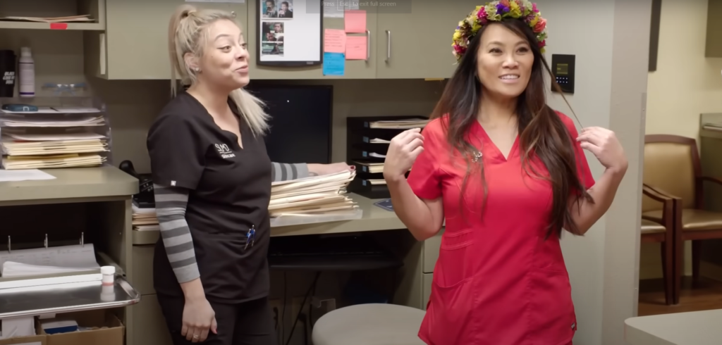 Dr Pimple Popper wearing a flower crown next to employee at skin clinic admin room 