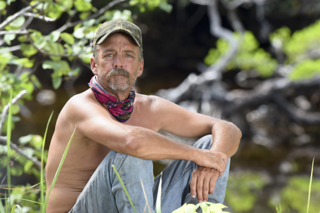 Keith Nale looks at camera wearing jeans and no shirt and cap and sits with his hands held over his knees on Survivor Cambodia: Second Chance