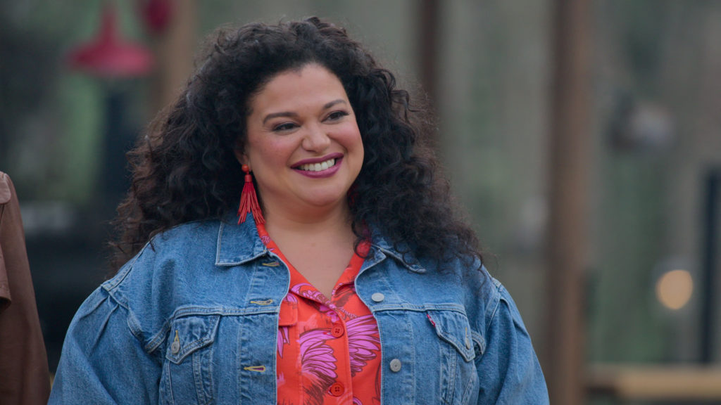 Michelle Buteau smiles wearing red and pink patterned shirt and blue denim jacket