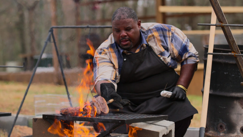 Thyron Mathews barbecues on open fire wearing plaid shirt and apron