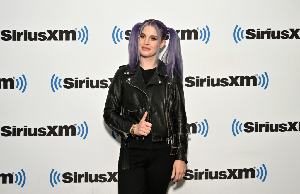 Kelly Osbourne poses smiling wearing black outfit and pigtails at SiruisXM event