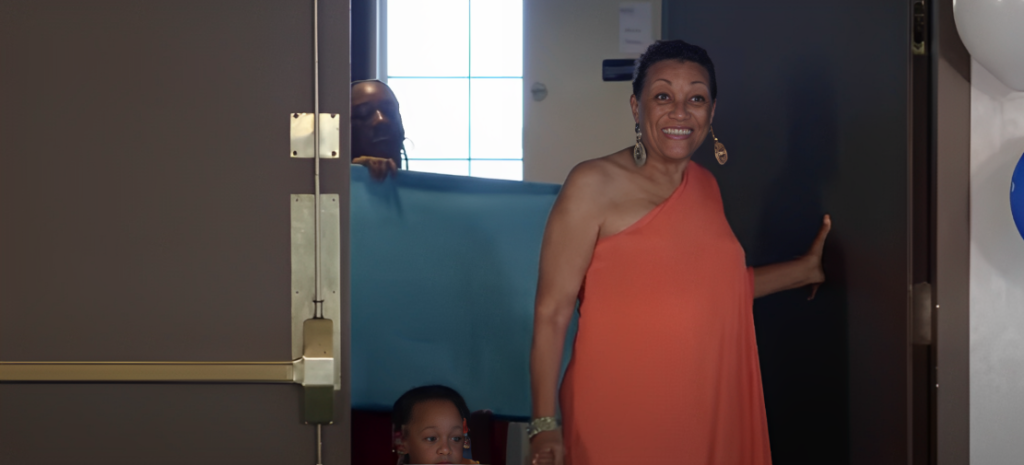 Marian Derrico smiles walking into room holding door open with one hand and holding child's hand in another wearing orange one-shoulder dress