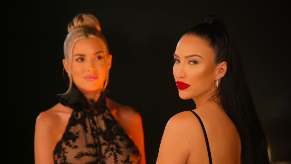 Heather Rae El Moussa stands next to Bre Tiesi as Bre looks at camera wearing red lipstick