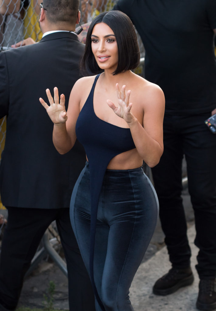 Kim Kardashian waves with both hands wearing one shoulder crop top and trousers and short dark brown bob