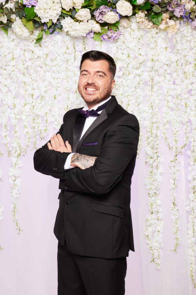 Luke smiles with right shoulder forward with arms crossed in a suit and purple bow tie, floral background behind. Seen down to knee level in black suit.