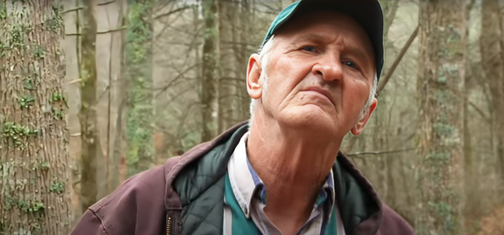 Jim Tom Hendrick speaks to camera filming Moonshiners wearing cap, shirt and jacket
