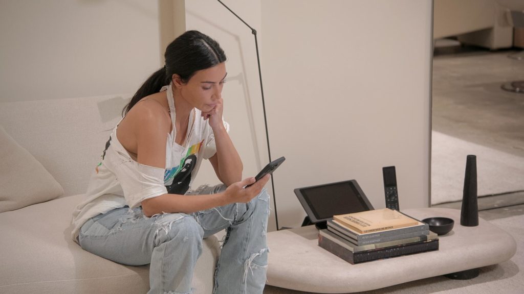 Kim Kardashian wears jeans and white top while sat on bed, looking at phone.