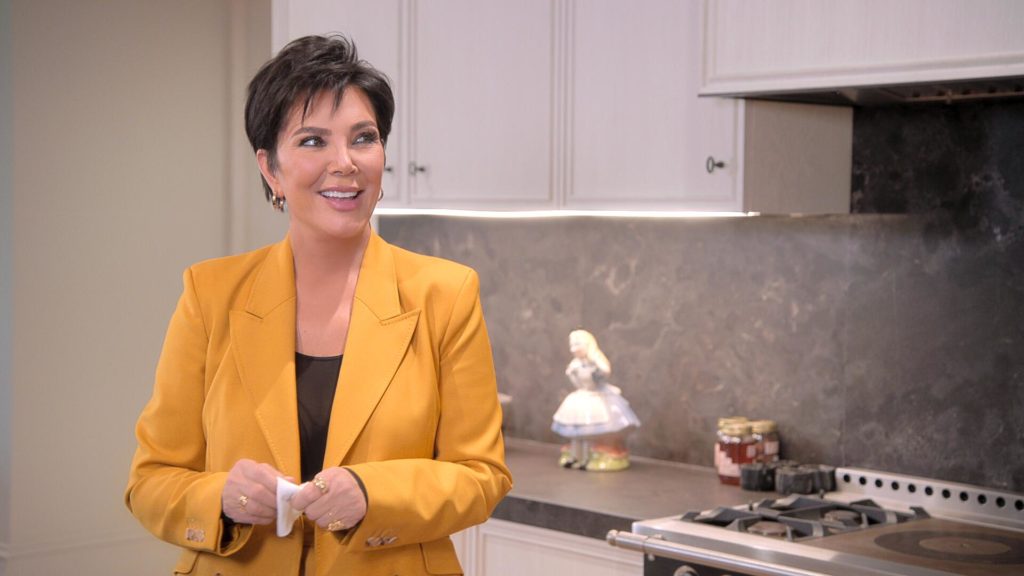 Kris Jenner smiles in kitchen wearing mustard color blazer and black top