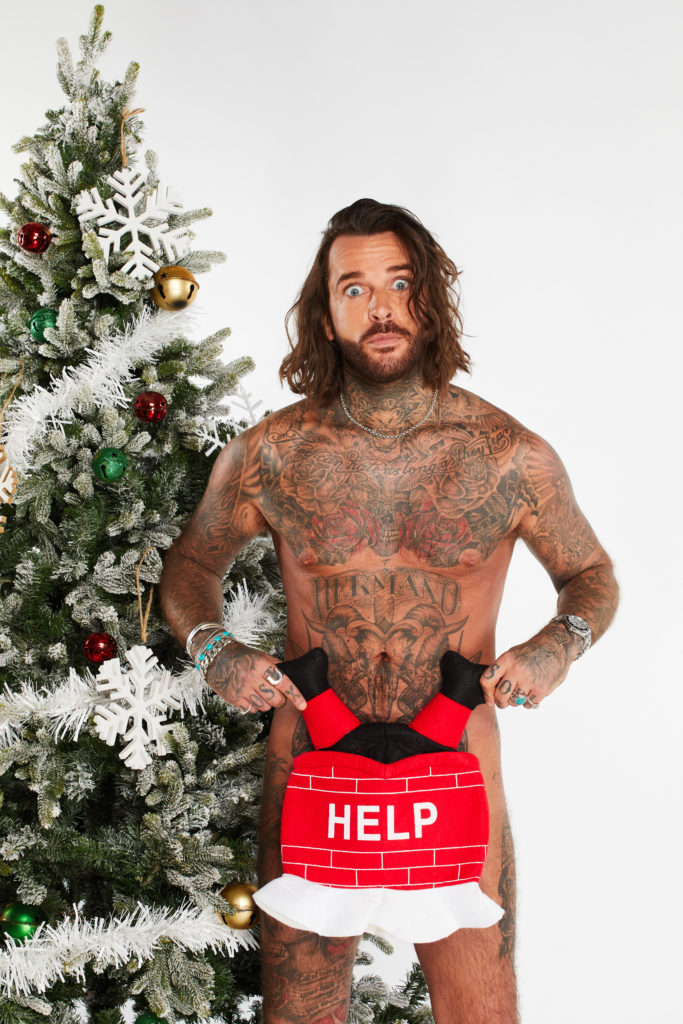 Pete Wicks poses naked next to Christmas tree as he holds red HELP upside down Santa toy over his private parts, as his body shows tattoos all over, with shocked look on face.